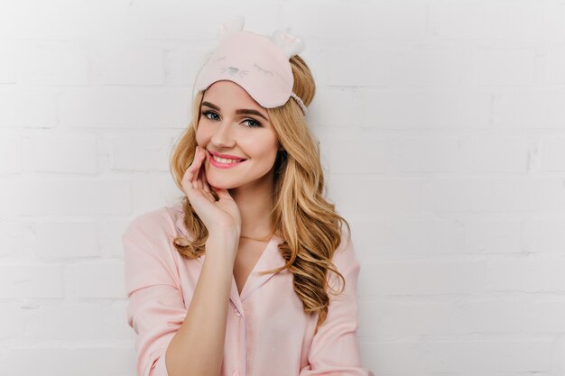 귀여운 수면 마스크에 매력적인 금발 소녀의 실내 초상화. 멋진 젊은 여성이 흰 벽에 포즈를 취하는 분홍색 잠옷을 입습니다.