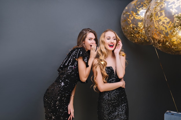 Indoor portrait of glamorous girls sharing rumors during party. Stunning ladies in black dresses talking about secrets while celebrating something with balloons.