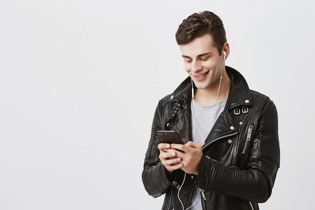 Indoor portrait of handsome caucasian guy in trende leather jacket holding mobile phone, messaging with his friends, telling them about his journey abroad having pleasant smile