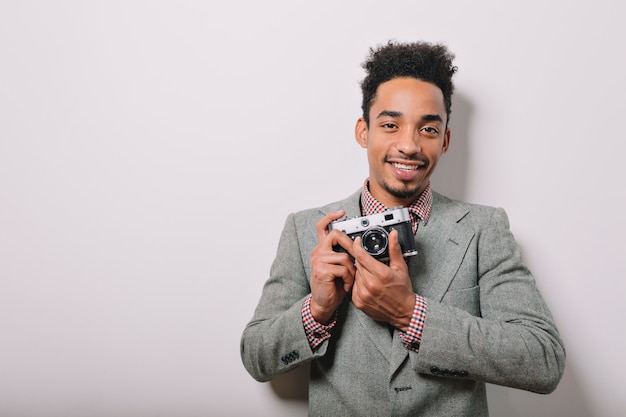 Indoor portrait of happy Afro-American male dressed in grey jacket holding camera in the hands on gray