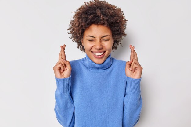 Indoor portrait of happy curly haired woman keeps fingers crossed believes in good luck prays for good exam results smiles gladfully wears casual blue turtleneck isolated over white background