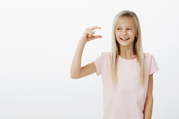 Indoor portrait of happy satisfied adorable young girl with fair hair, looking at tiny thing, shaping it with fingers