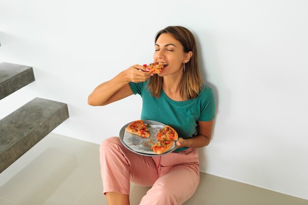 Indoor portrait of happy woman eating pizza with cheese, sitting on the flor in modern house