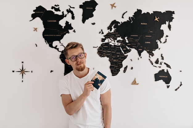 Indoor portrait of happy young european man with passport posing  over world map. Preparing for travelling, vacation trip.