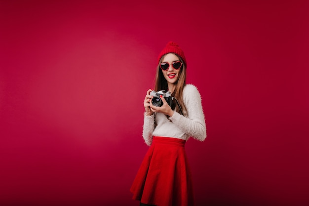 Ritratto dell'interno della ragazza interessata in occhiali da sole e cappello rosso isolato sullo spazio bordeaux