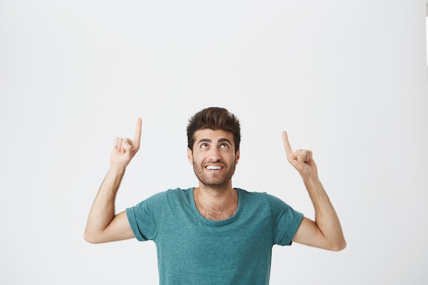 Крытый портрет радостного бородатого испанского парня с довольным выражением лица, одетый в синюю футболку, смеясь и указывая вверх ногами на белой стене. Копировать пространство