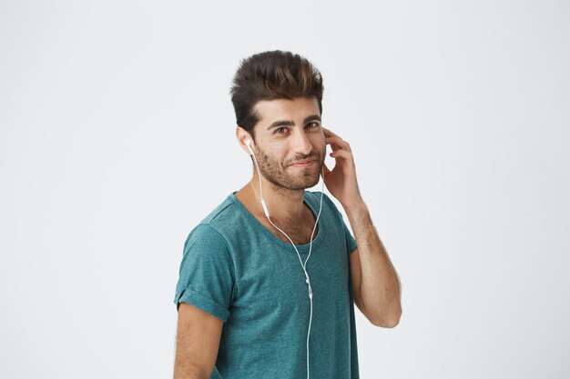 Indoor portrait of joyful spanish man smiling gently in blue t-shirt, with trendy hairstyle and beard, looking  listening his fave music.