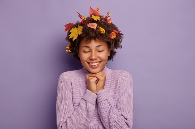 素敵な幸せな女性の屋内の肖像画は、両手をあごの下に保ち、暖かく乾燥した秋の天気を楽しみ、喜びを感じ、髪に葉とナナカマドの果実を持ち、セーターを着て、紫色の壁に隔離されています