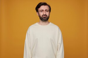 Indoor portrait of man wears white sweatshirt posing over yellow background starring into camera with negative sad facial expression