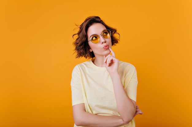 Indoor portrait of pensive brunette girl in light-yellow t-shirt. Glad short-haired woman in sunglasses looking up and thinking about something.