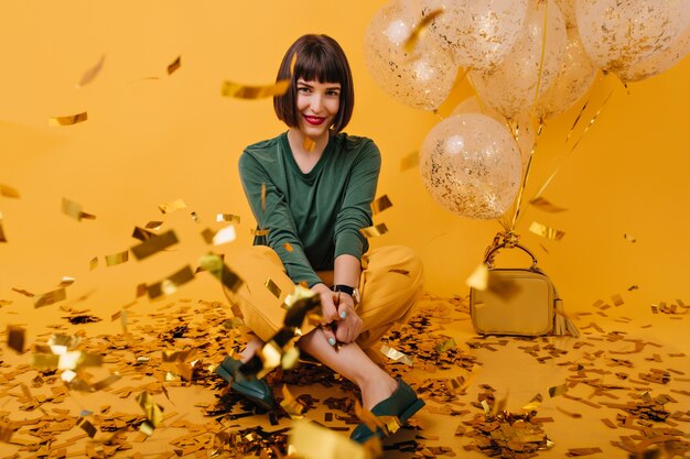 Indoor portrait of pleasant brunette girl sitting on confetti. cheerful european lady in green sweater posing.