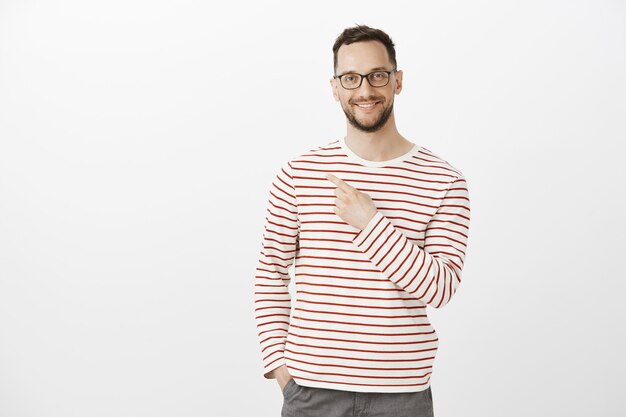 Indoor portrait of pleased good-looking man model in glasses, smiling broadly and pointing back or at upper left corner