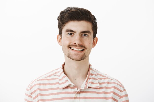 Indoor portrait of pleased handsome ordinary european man with moustache and beard, smiling broadly, feeling confident and happy while making arrangement for wedding over gray wall