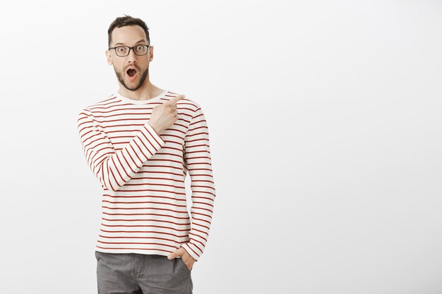 Indoor portrait of pleased impressed handsome man in eyewear, pointing at upper right corner, dropping jaw from positive emotions