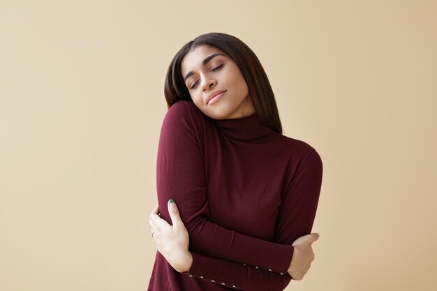 Indoor portrait of positive relaxed young mixed race brunette woman closing eyes with pleasure, keeping arms around herself, enjoying soft fabric of her new maroon cashmere turtleneck sweater