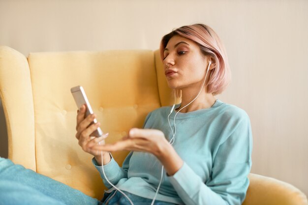 Indoor portrait of pretty girl with nose ring wearing earbuds while talking to friend using webcam on cell phone