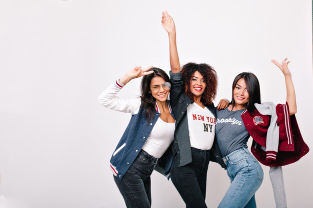 Indoor portrait of pretty international friends funny dancing and waving hands. Adorable asian girl in blue jeans spending weekend with university mates and posing.