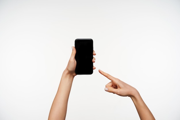 Indoor portrait of pretty young female's hands holding mobile phone and showing on black screen with forefinger, being isolated on white