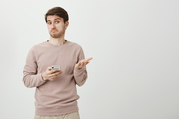 Indoor portrait of puzzled and clueless young man gesturing and shrugging while holding smartphone and looking confused