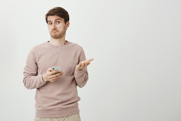 Портрет озадаченного и невежественного молодого человека в помещении, жестикулирующего и пожимающего плечами, смущенно держа смартфон в руке