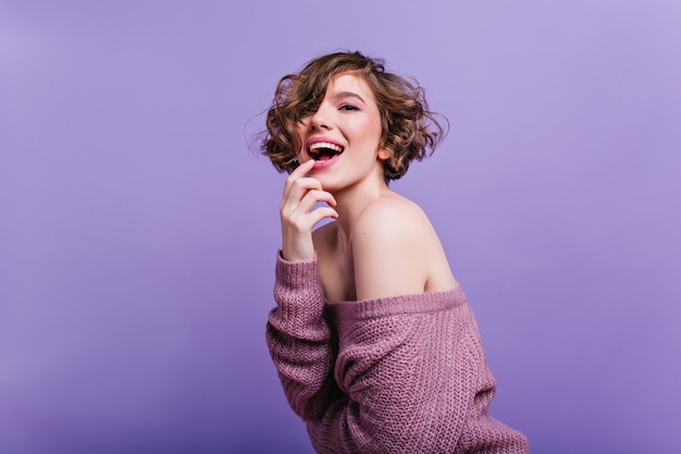 Indoor portrait of sensual short-haired woman in knitted attire posing  Playful curly brunette girl in sweater standing on purple wall.