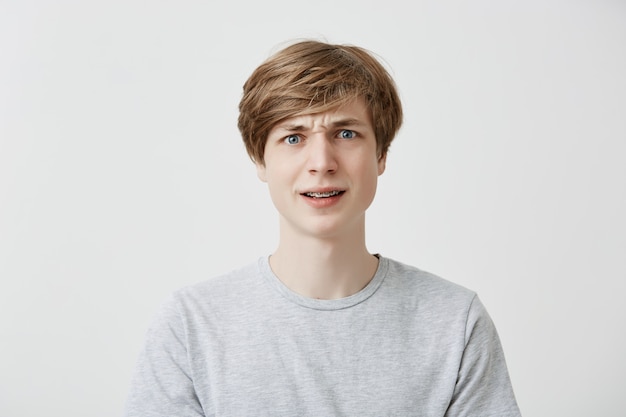 Indoor portrait of serious dissatisfied puzzled emotional fair-haired young man with braces, frowns his face in bewilderment, reacts on teachers`s remark. Peope and reaction concept