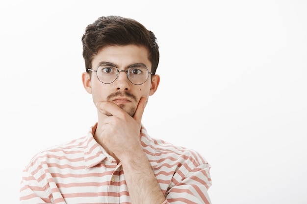 Free photo indoor portrait of serious focused male nerd in trendy round glasses, rubbing chin with hand and staring, thinking or making decision, solving mathematical issue over gray wall