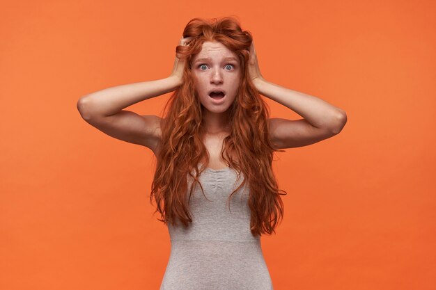 Indoor portrait of shocked attractive young redhead woman with wavy lond hair standing over orange background, clutching her head with wide mouth opened and rounding eyes