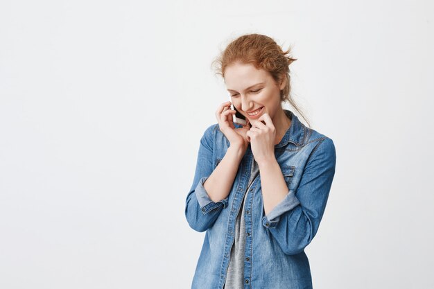Indoor portrait of shy cute college student with ginger hair combed in bun talking on smartphone and being confused or embarassed speaking with guy he likes