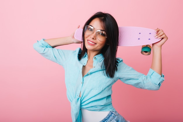 Ritratto dell'interno della donna asiatica spettacolare in camicia di cotone e occhiali da sole. modello femminile latino dai capelli neri con longboard sorridente in camera rosa.