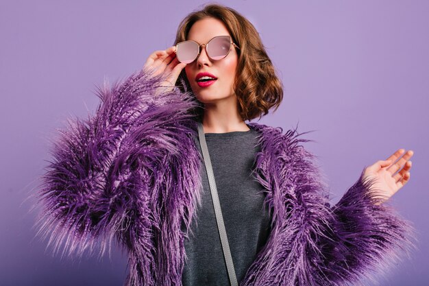 Крытый портрет стильной европейской женской модели в очках позирует на фиолетовом фоне
