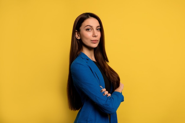 Indoor portrait of successful young woman with long dark hair wearing blue jacket posing with folded arms over yellow wall