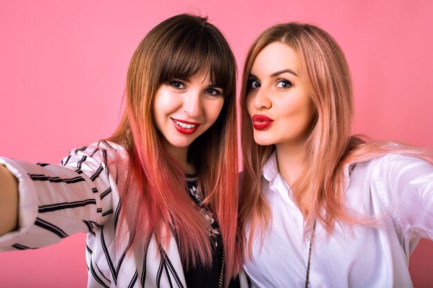 Indoor  portrait of two happy best friends sisters women, wearing trendy black and white clothes and pink hairs, making selfie, enjoying time together