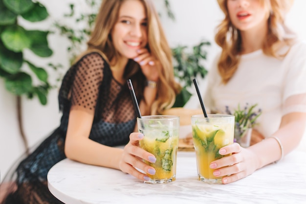 Indoor portrait of two joyful girls chilling in cafe with glasses of tasty cocktails on foreground