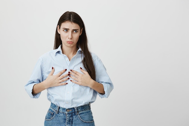 Indoor portrait of unsatisfied and sad girl with gloomy smile and jealous look holding her breast and looking aside standing over gray background Woman wishes she had at least cup B