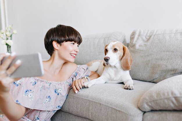 Indoor portrait of wonderful dark-haired girl making selfie with beagle dog lying on sofa