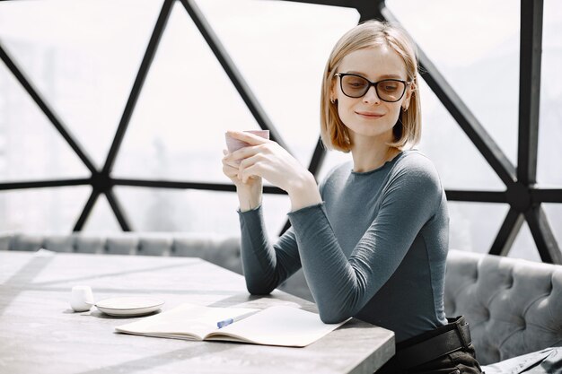 Indoor portrait of a young businesswoman sitting in a cafe and drinking a coffee. Blonde girl wearing sunglasses and jacket