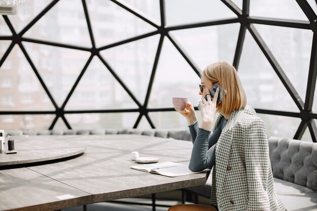 電話で話し、コーヒーを飲むカフェに座っている若い実業家の屋内の肖像画。サングラスとジャケットを着ているブロンドの女の子