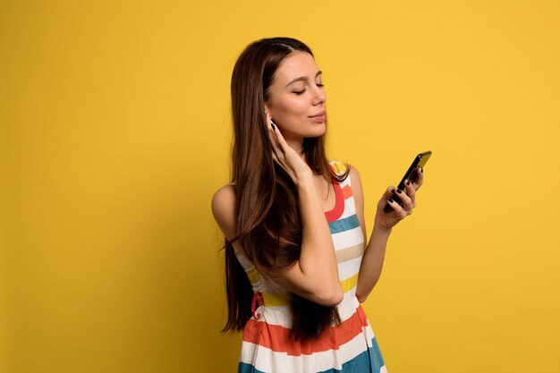 Indoor portrait of young charming girl with long dark hair is listening music and looking at phone over yellow wall