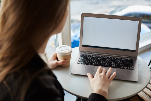 Indoor rearview of stylish woman sitting in cafe, drinking cup of coffee and working with laptop