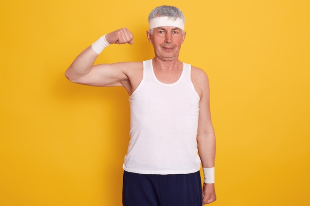Indoor senior man wearing sport clothing and headband, standing with one hand up and showing his biceps, being photographed after doing physical exercises. Healthy lifestyle concept.