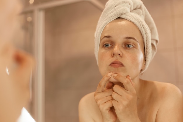 Indoor shoot of young adult beautiful woman wearing bath towel standing in bathroom and looking for or squeezing acne on chin, mirror reflection, beauty procedures.