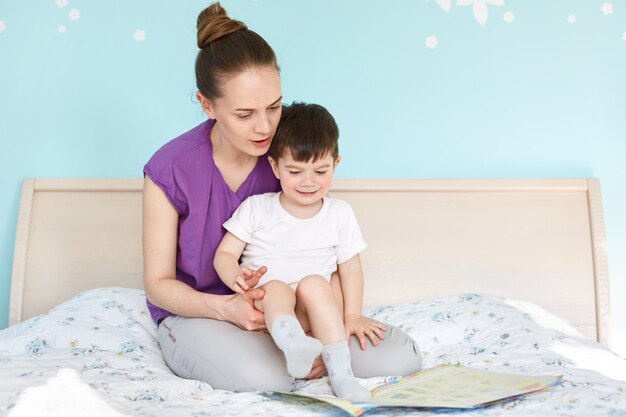 Indoor shot of affectionate young mother holds and embraces her small son, look attentively at book with colourful pictures