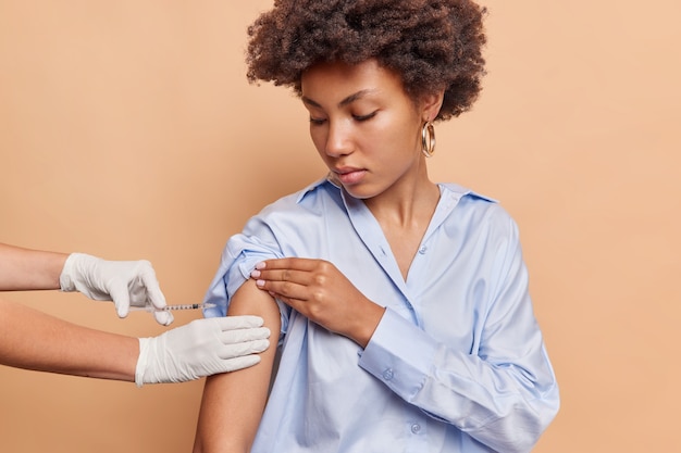 Il colpo al coperto della donna afroamericana ottiene la vaccinazione in braccio per la salute immunitaria impedisce di essere infettato dal coronavirus indossa una camicia blu isolata sul muro beige