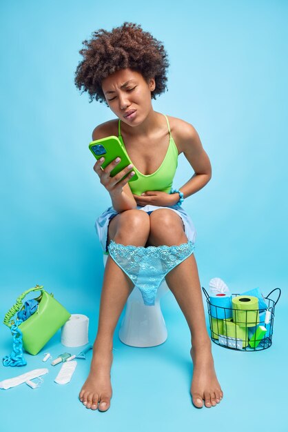Снимок афроамериканки, страдающей спазмами в животе, в помещении: запор искал в интернете, какое лекарство принять, держит мобильный телефон в туалете на унитазе