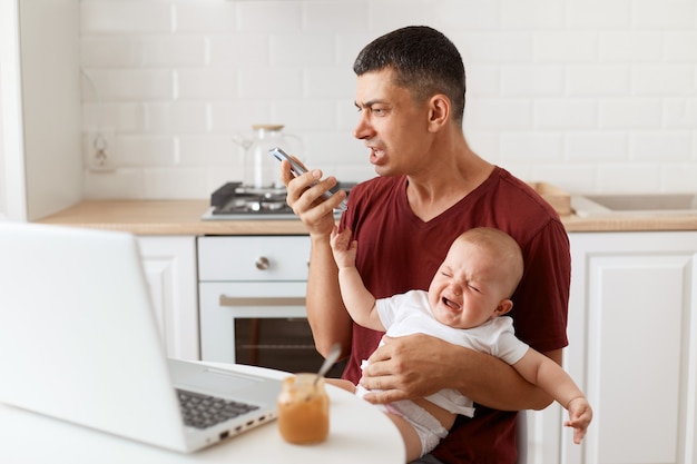 Крытый снимок агрессивного мужчины-брюнетки в темно-бордовой футболке в повседневном стиле, сидящего за столом на кухне со своей младенческой дочерью, кричащего во время записи голосового сообщения.