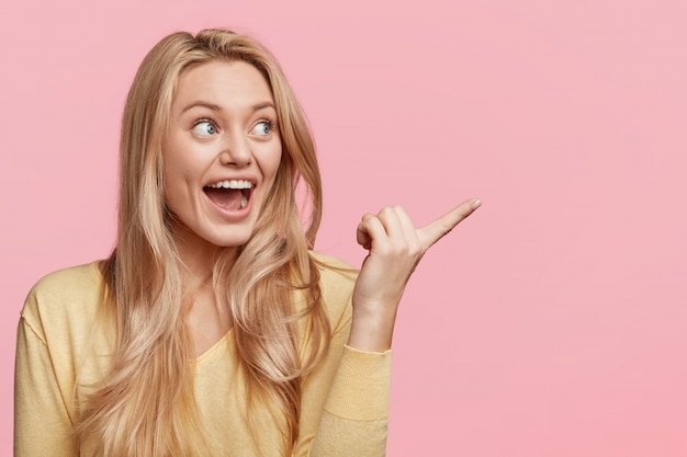 Free photo indoor shot of amazed blonde woman has overjoyed expression, indicates with excited look aside, poses against pink wall with blank copy space for your advertising content. wow, that`s great!