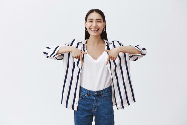 Indoor shot of amazed carefree beautiful woman in striped blouse and jeans, laughing out loud from happiness, pointing down with index fingers, showing interesting and amusing thing over grey wall