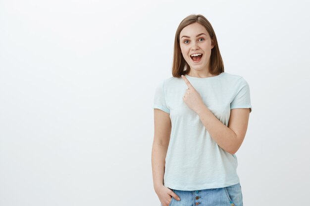 Indoor shot of amazed energized and optimistic cute woman in trendy t-shirt smiling cheerfully pointing at upper left corner with index finger near chest enjoying spending time fun and amused