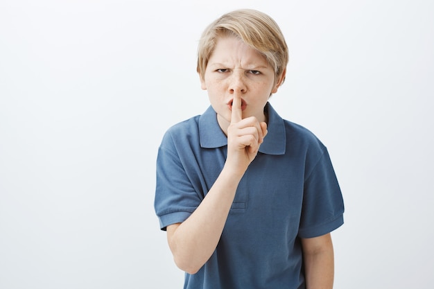 Indoor shot of angry annoyed blond brother in casual t-shirt, frowning, saying shh while making shush gesture with index finger over mouth, demanding to stay quiet